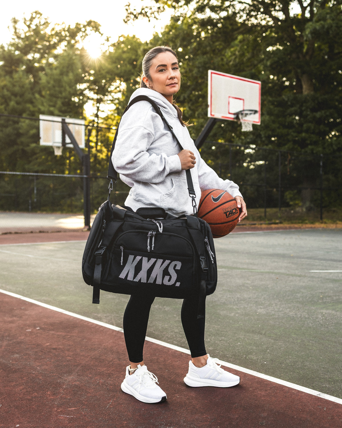 Woman carrying a black KXKS basketball duffle bag over her shoulder while holding a basketball on an outdoor court