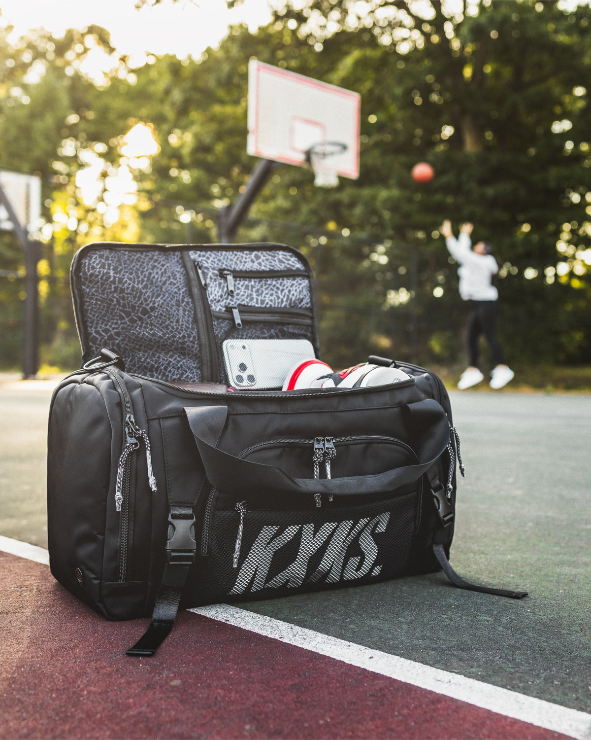 Black KXKS basketball gym bag open on a court with sneakers and phone visible, and a player shooting hoops behind
