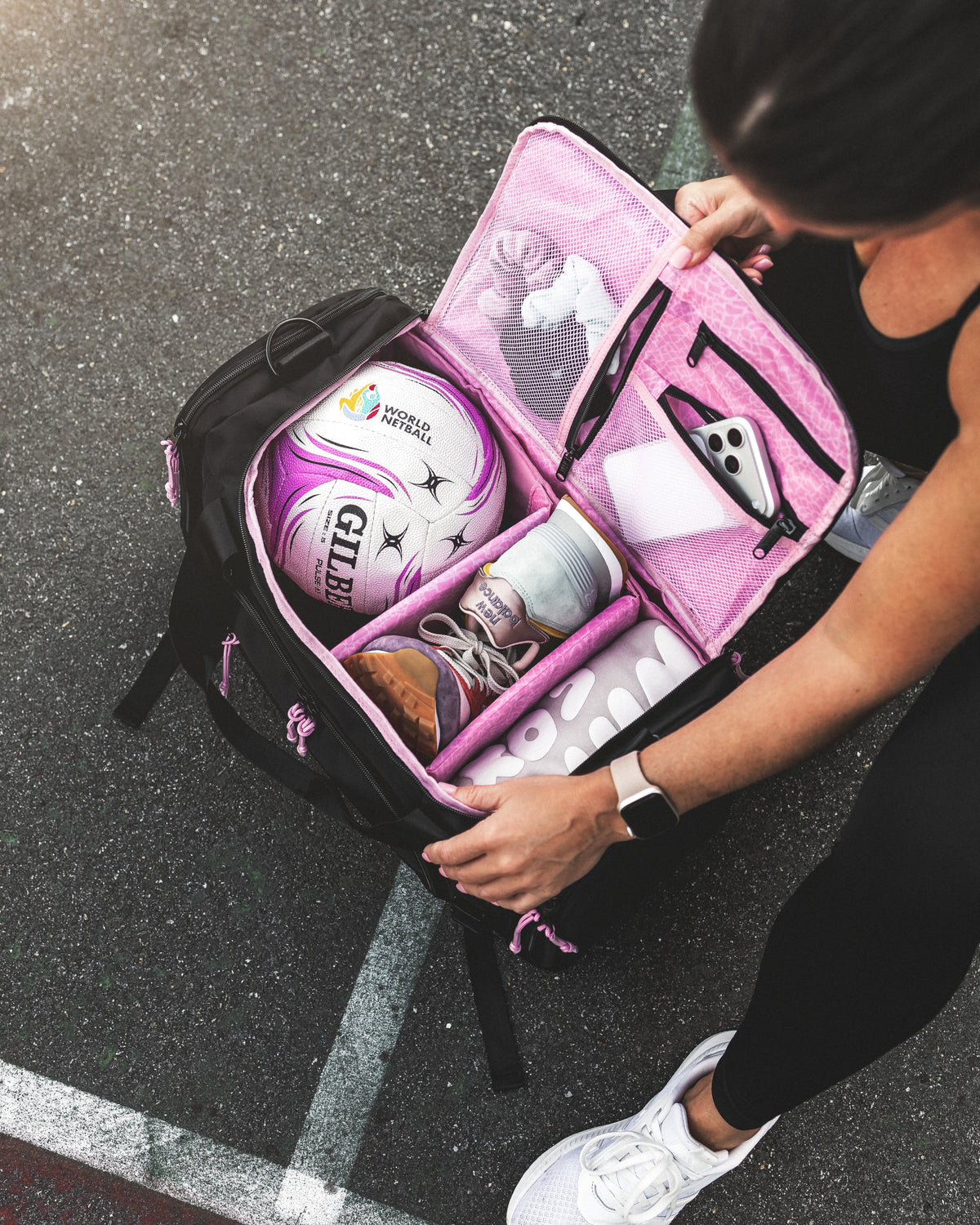 Open black netball bag with pink lining holding a netball, sneakers in a separate shoe compartment and a phone in the mesh pocket