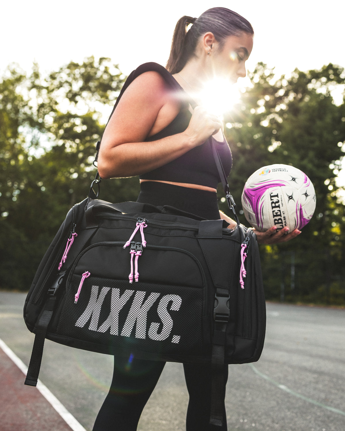 Woman with a black KXKS netball duffle bag with pink zip cords on her shoulder, holding a netball in the sun