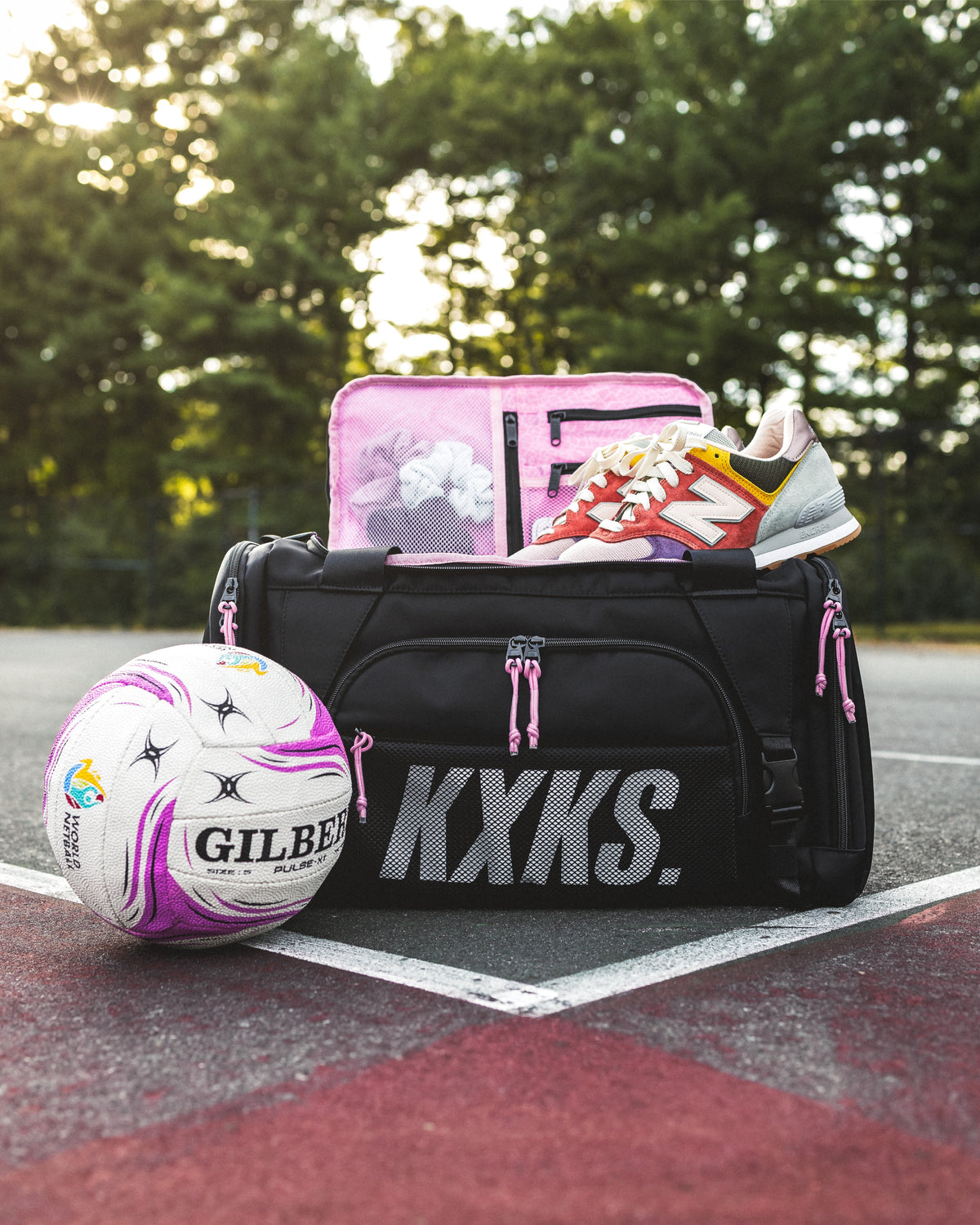 KXKS netball duffle bag with pink lining open, a netball on the court beside it and sneakers resting on top