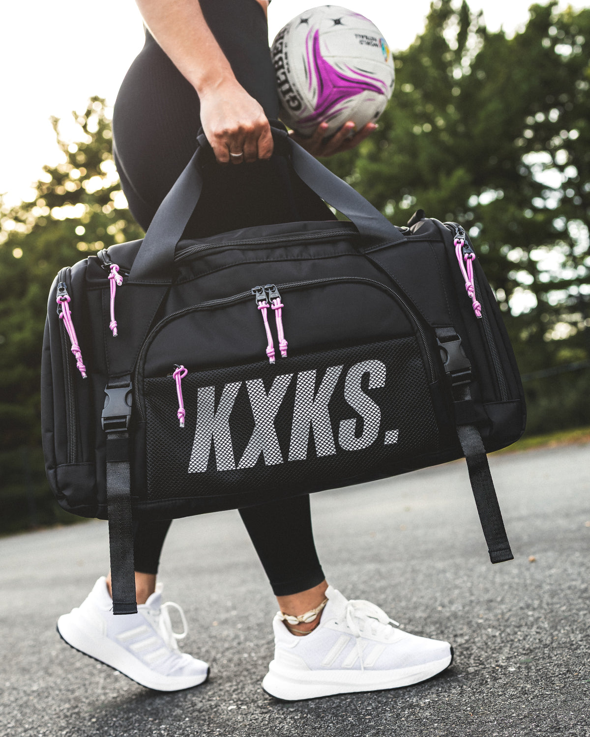 Black KXKS netball sports bag with pink zip cords carried by the top handle, with a netball in her other hand