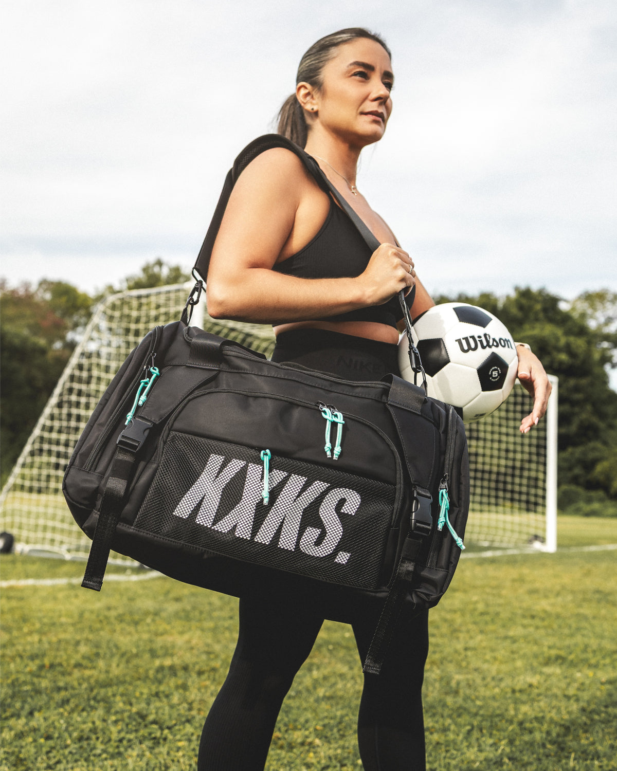 Woman carrying a black KXKS soccer duffle bag with teal zip cords over her shoulder in front of a goal