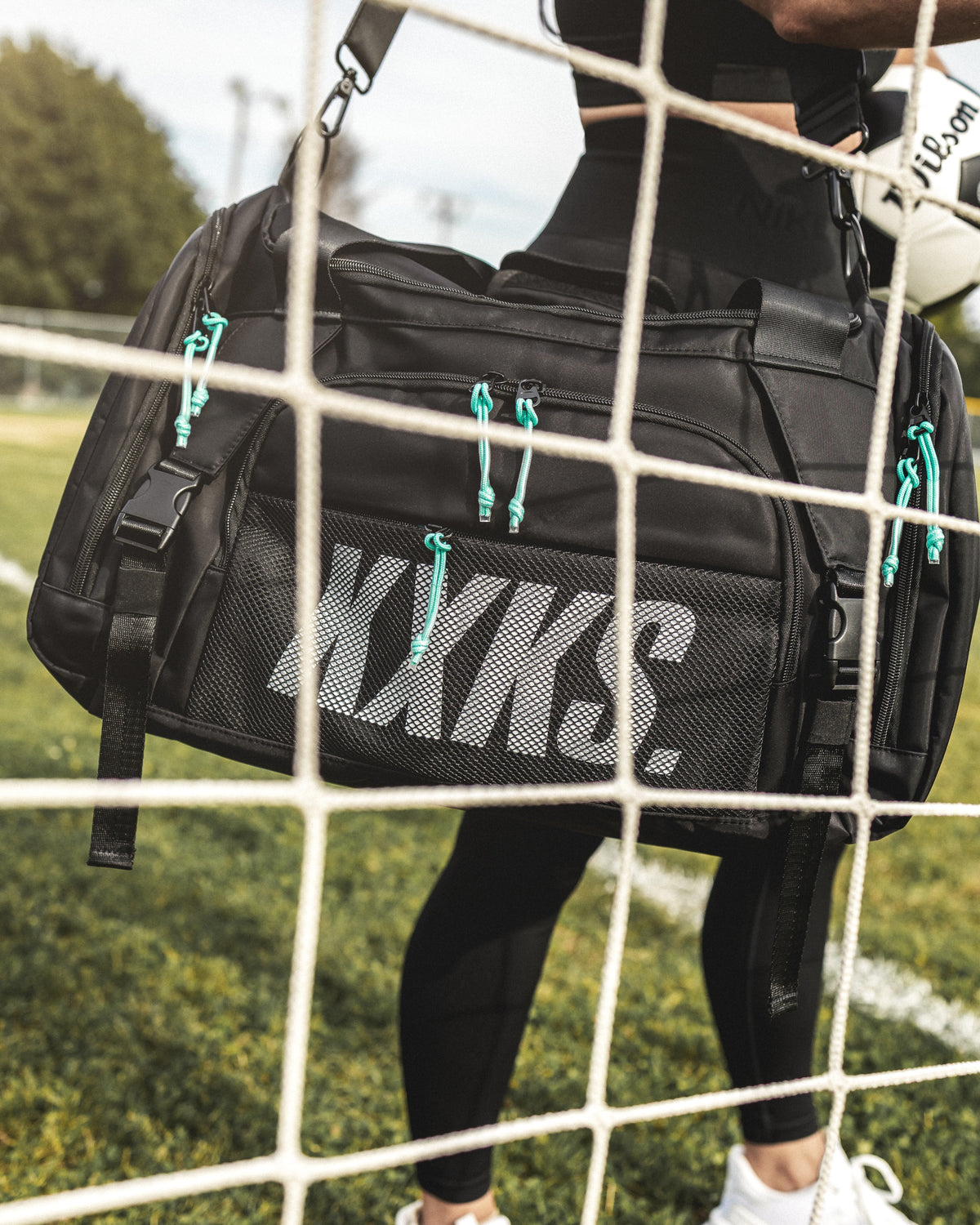 Close-up of a black KXKS soccer gym bag with teal zip cords and a mesh front pocket, seen through a goal net